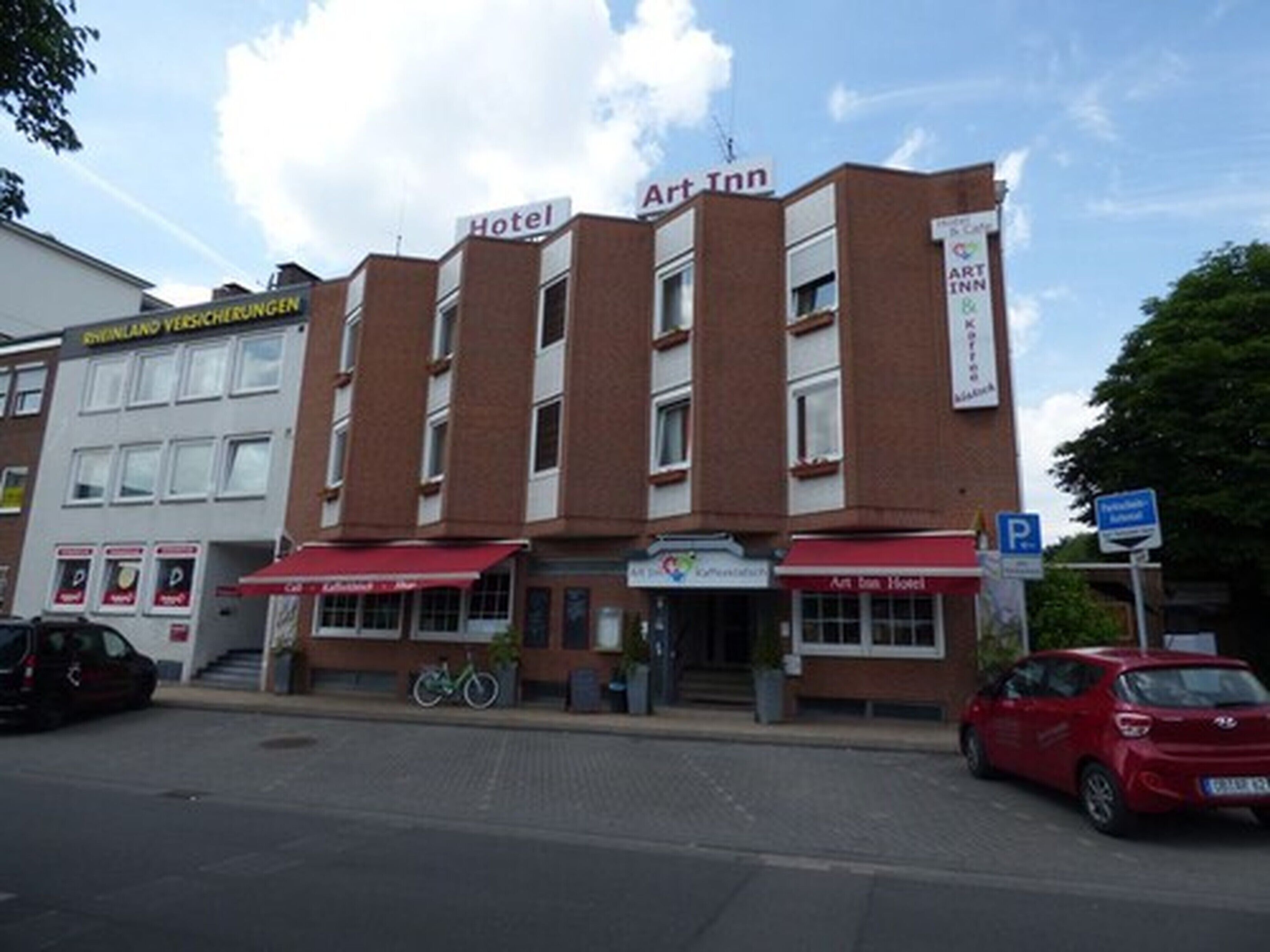 Un hôtel avec un auvent rouge et une enseigne indiquant « Art Inn ». Un vélo est garé devant, et une voiture rouge est stationnée dans la rue.