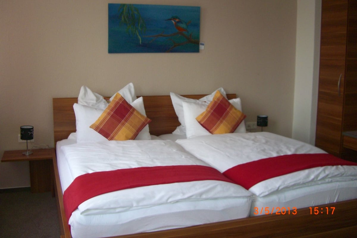 A double bed with white linens, two red and yellow plaid pillows, and a red throw blanket. There is a painting of a bird on the wall above the bed.
