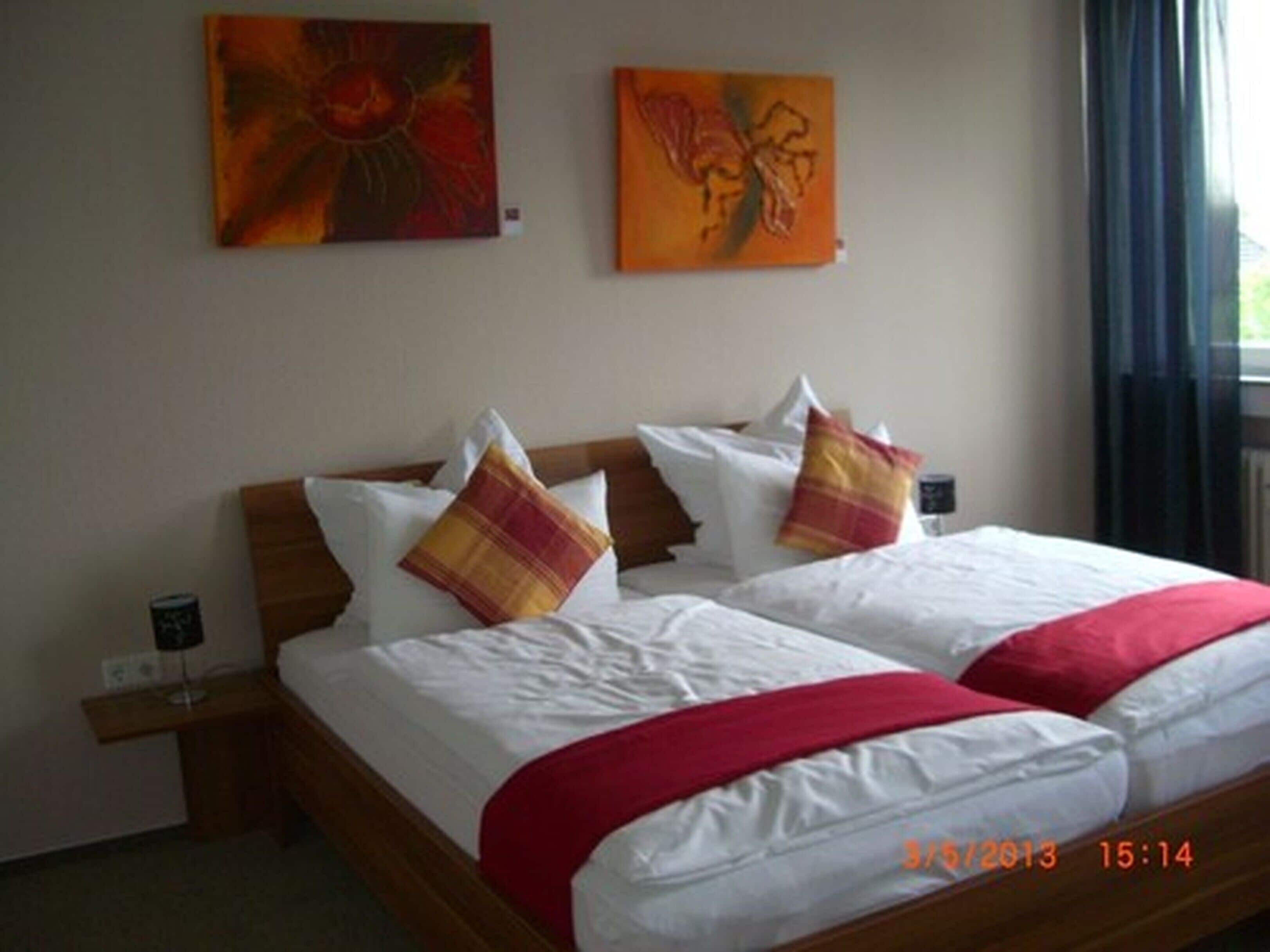 Una cama doble con ropa blanca y detalles rojos, dos almohadas, una mesita de noche y dos cuadros en la pared.