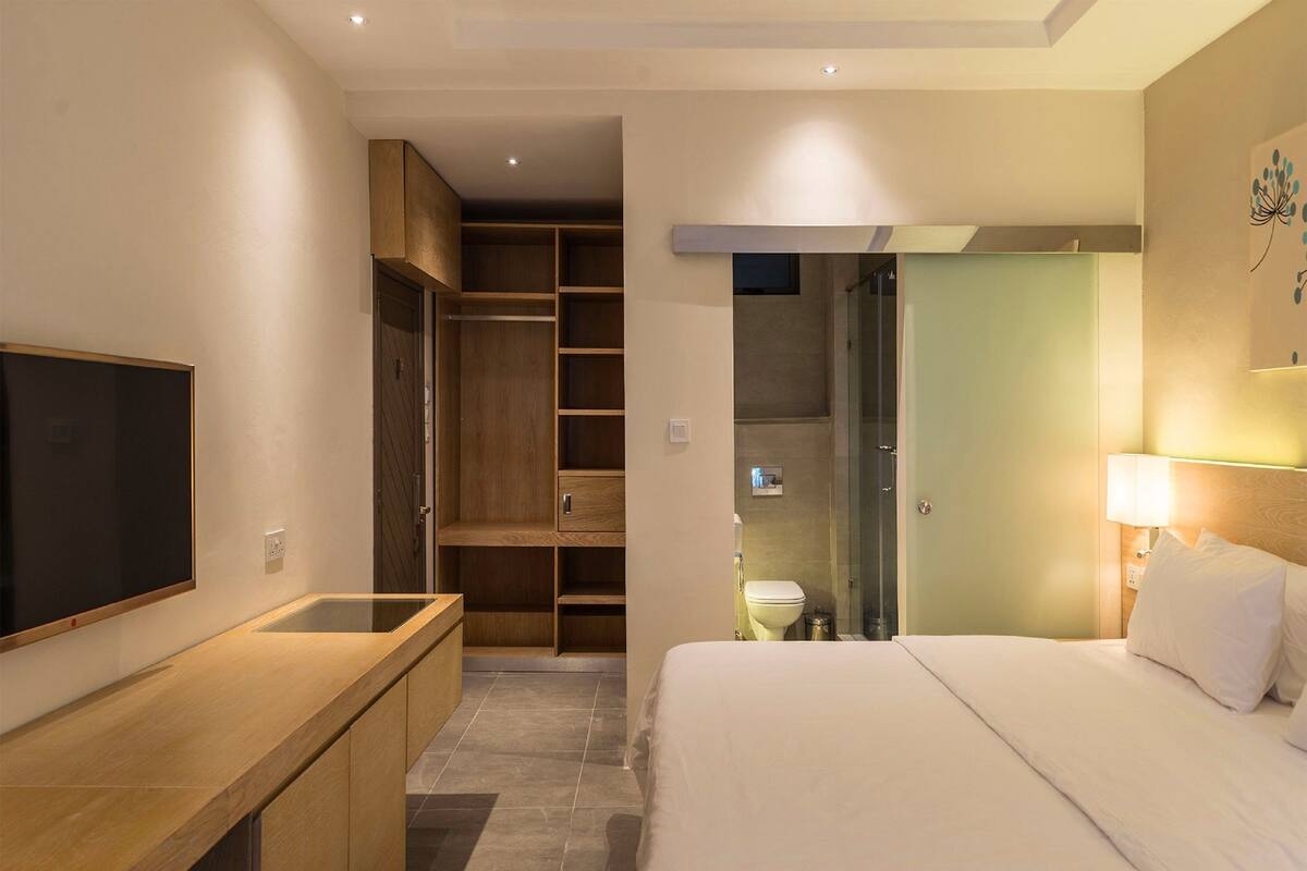 A hotel room with a bed, a TV, a wooden desk, a wardrobe, and a bathroom visible through an open door.