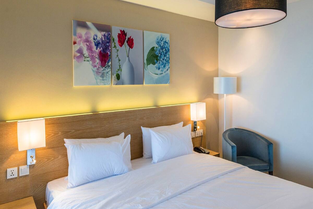 A hotel room with a large bed, two bedside lamps, a chair, and three wall-mounted paintings of flowers.