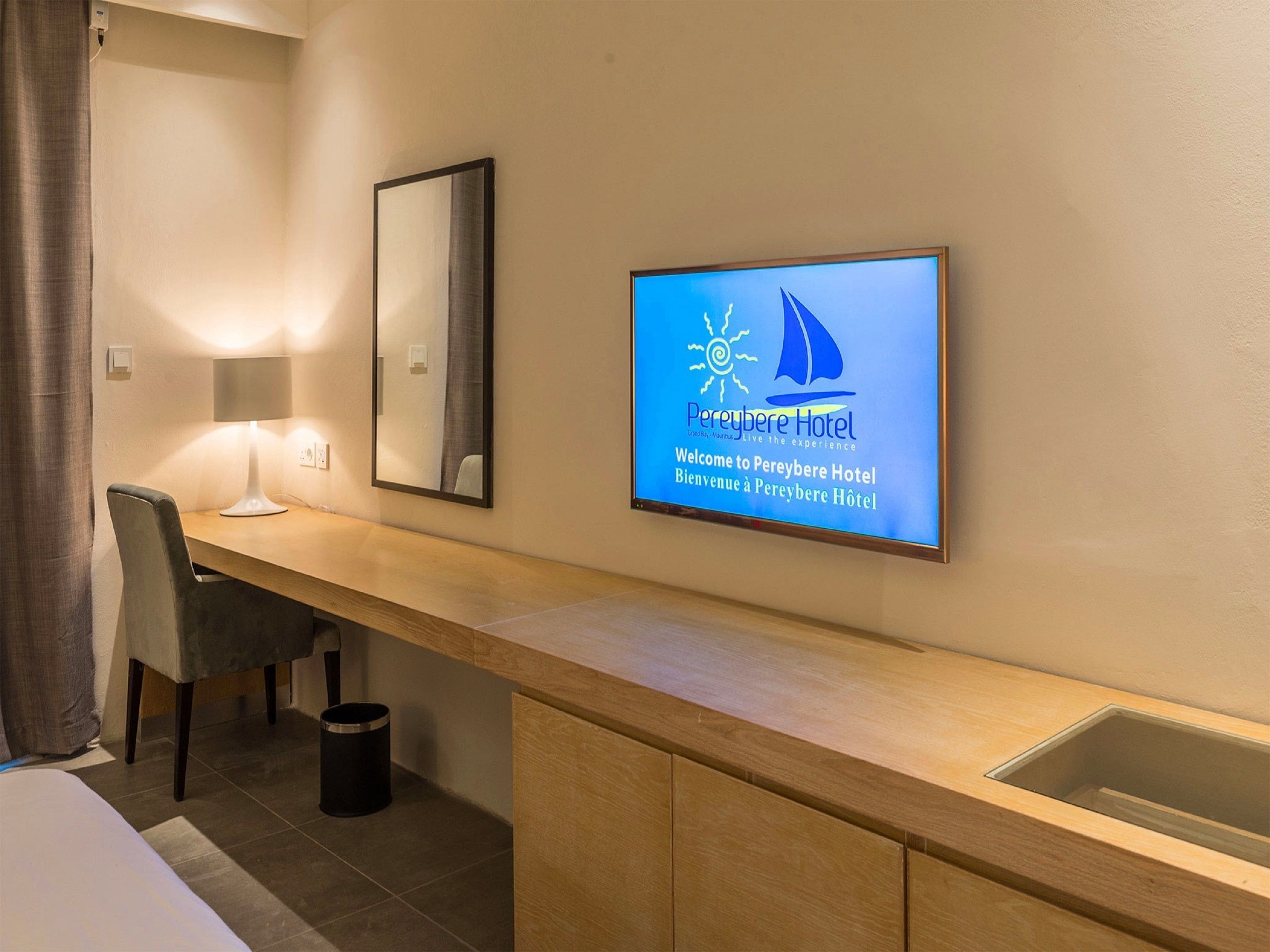 Camera d'albergo con una scrivania in legno, una sedia, una lampada, un cestino e una TV a parete che mostra il logo dell'hotel.