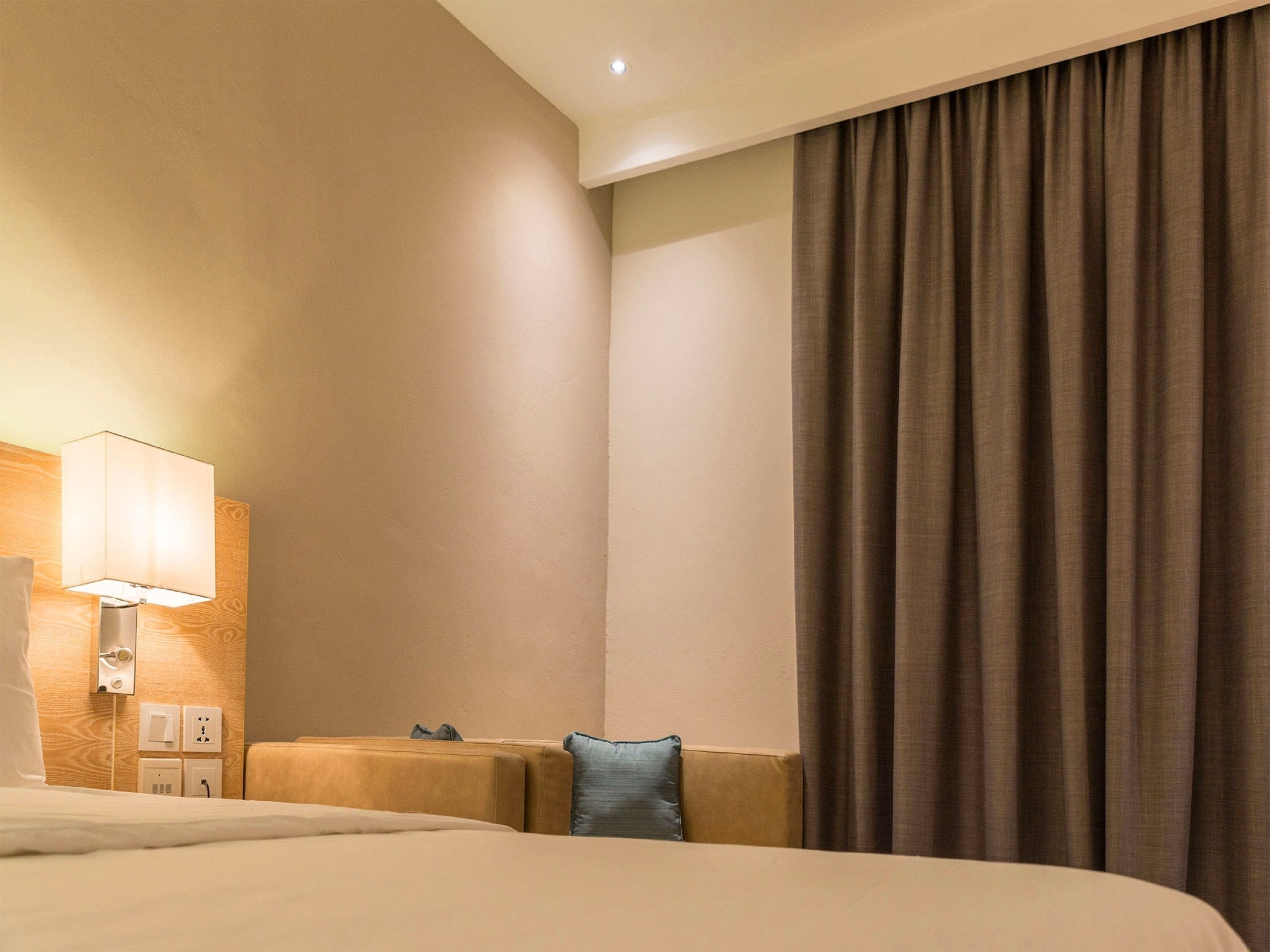 Una habitación de hotel con cama, lámpara de noche, mesita de noche y ventana con cortinas.