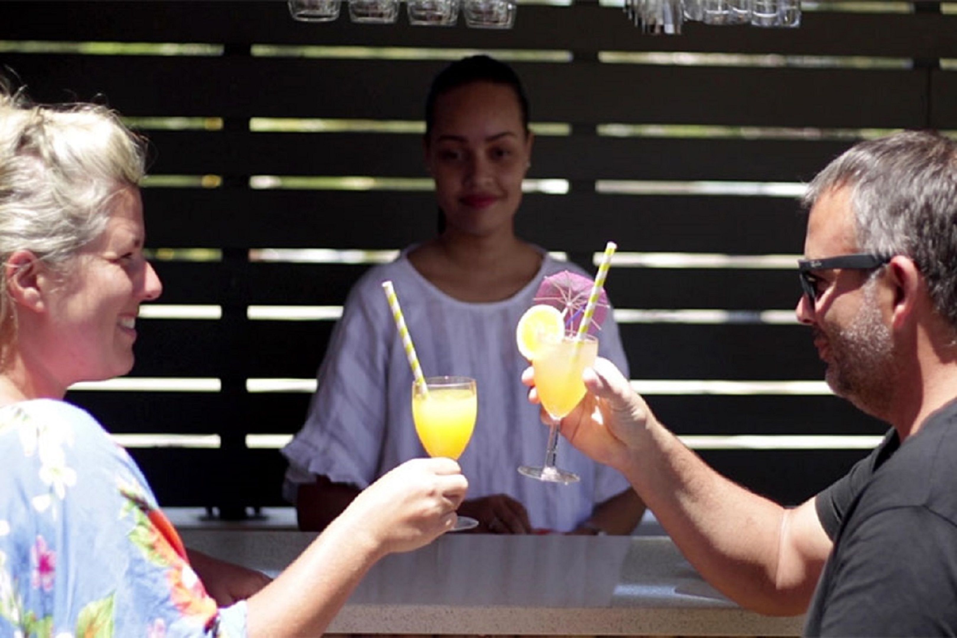 Un groupe de personnes trinquant avec des boissons dans un espace bar en plein air, doté d’un aménagement simple et fonctionnel et d’un décor en bois.