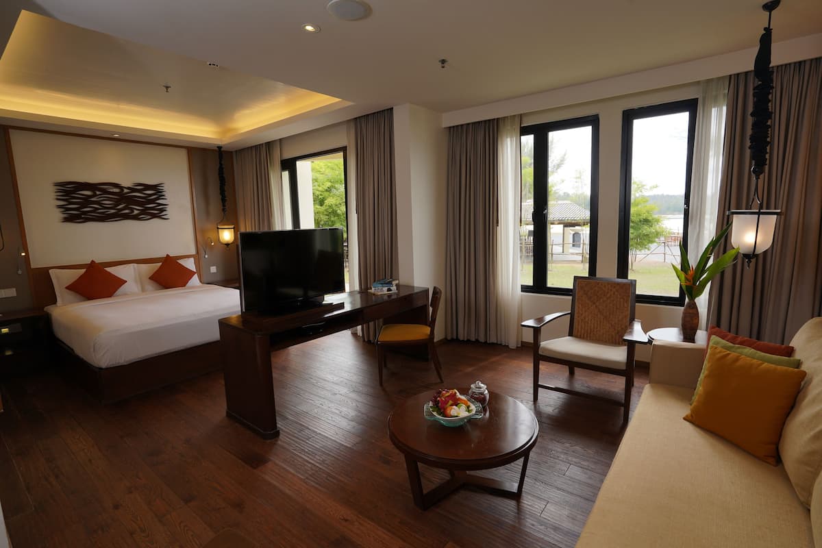 A hotel room with a bed, a desk with a TV, a sofa, a coffee table, and large windows with curtains.