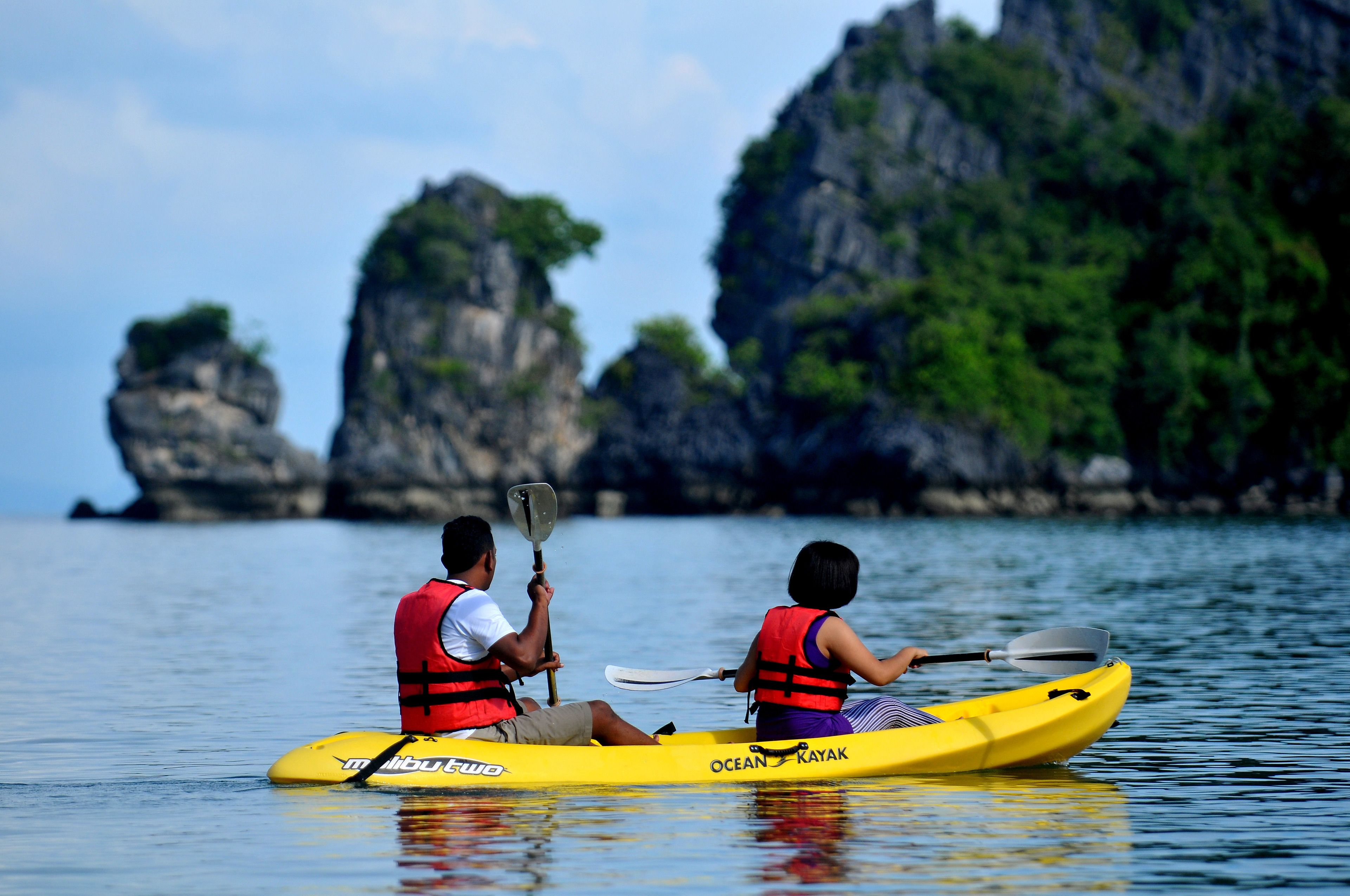 Two people kayaking on a calm sea with rocky cliffs in the background.