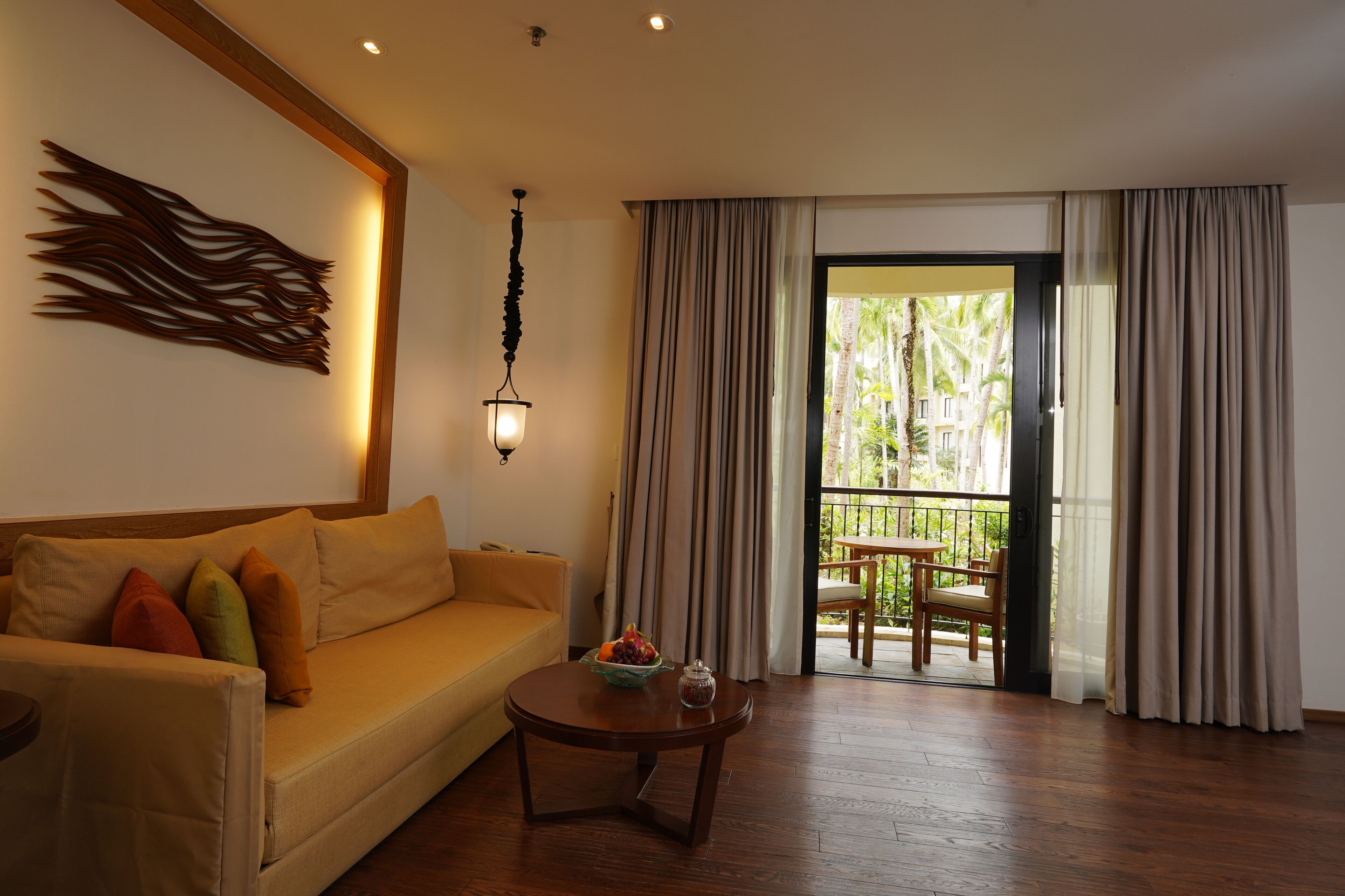 Una sala de estar con un sofá, una mesa redonda con un cuenco y un tarro, una obra de arte en la pared, y vistas a un balcón con una silla.