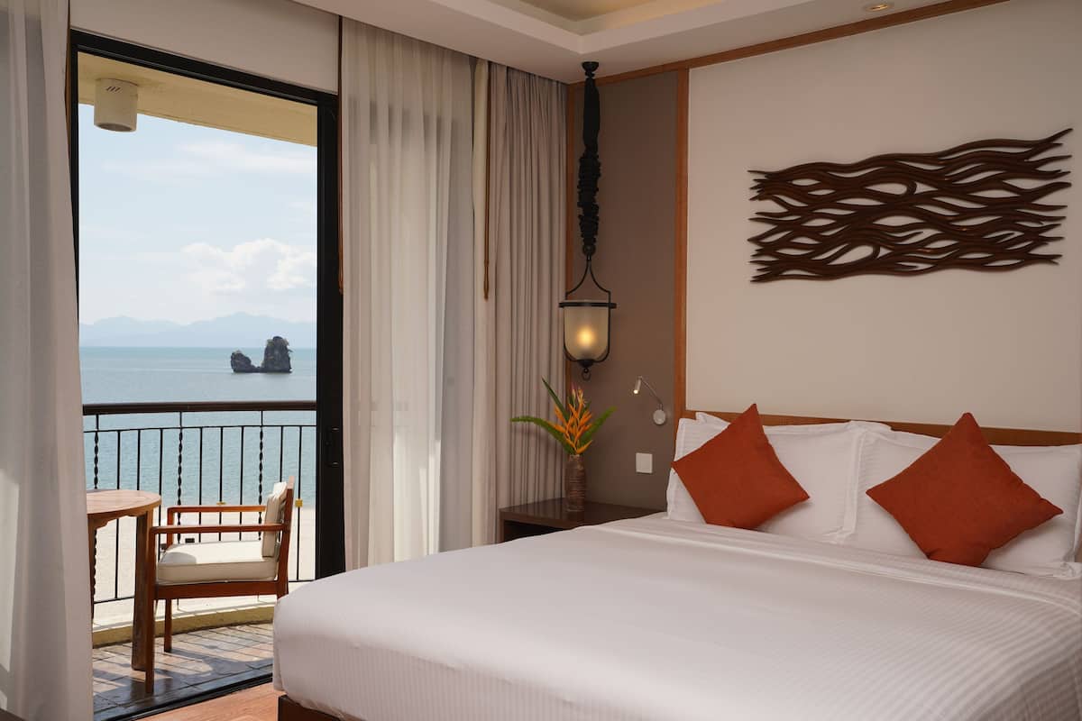 A hotel room with a bed, a nightstand, a lamp, a vase with a plant, a view of the sea, and a balcony with a chair.