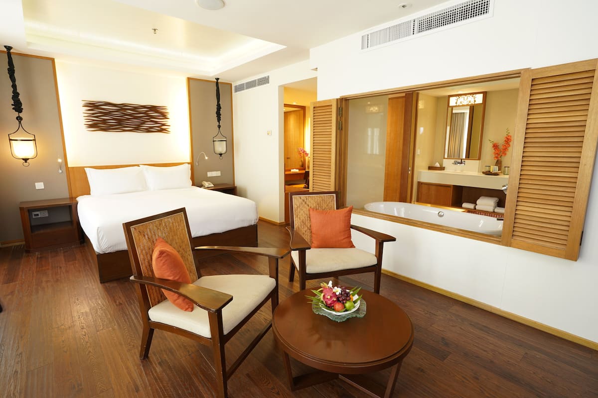 A hotel room with a bed, two chairs, a round table with fruit, a large mirror, and a bathroom with a bathtub.