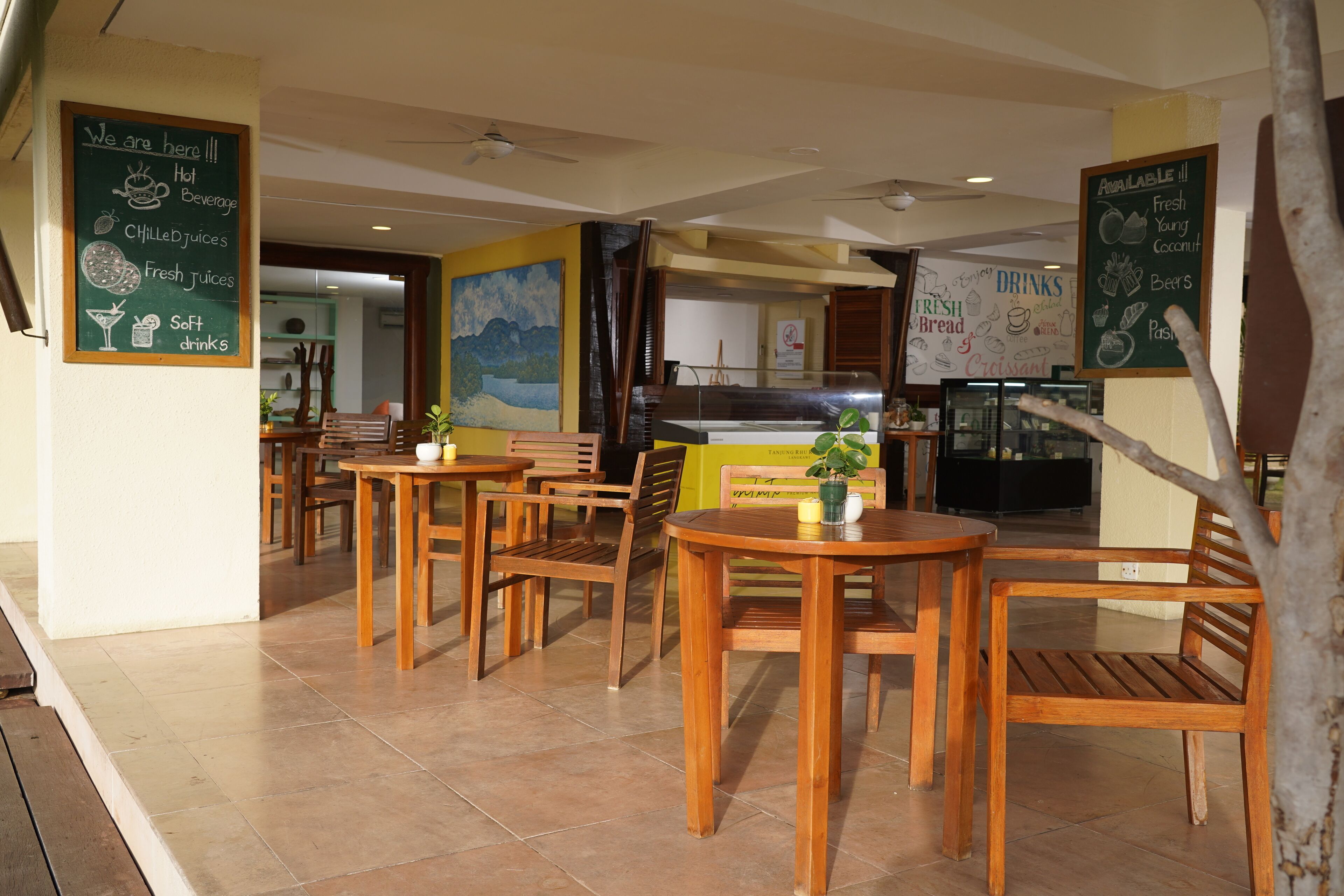 Un café avec des tables et des chaises en bois, un tableau noir servant de menu, et une vue sur la cuisine en arrière-plan.