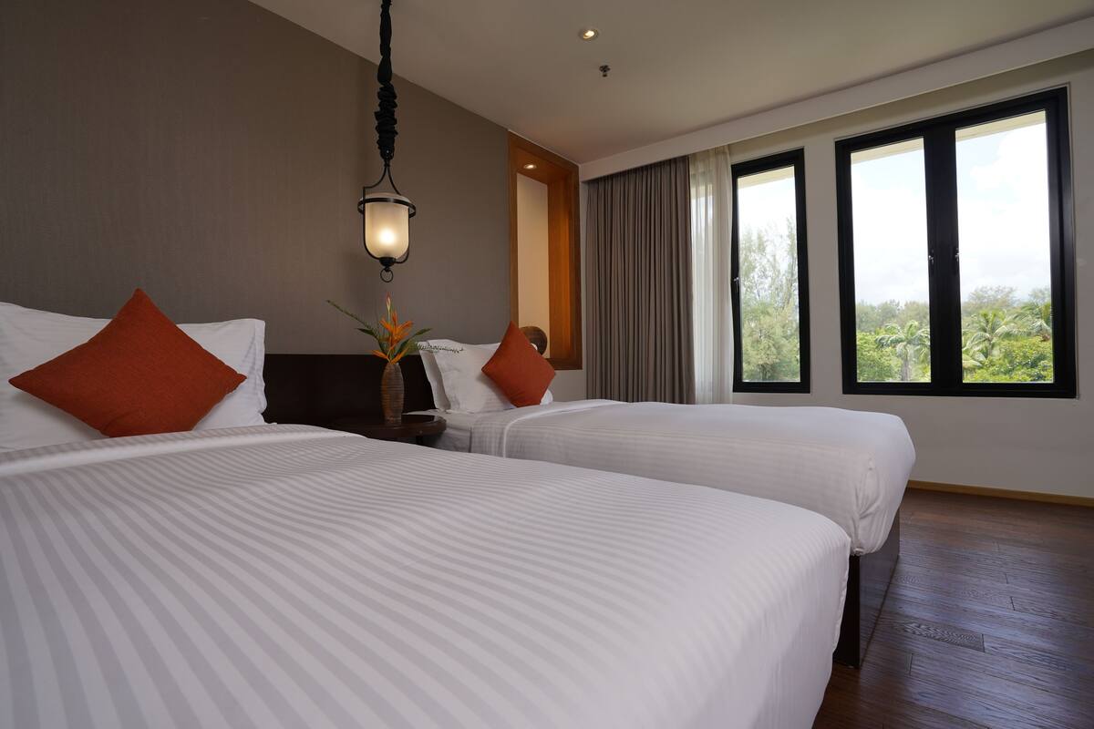 A hotel room with two beds, a nightstand, a lamp, and a large window with a view of greenery.