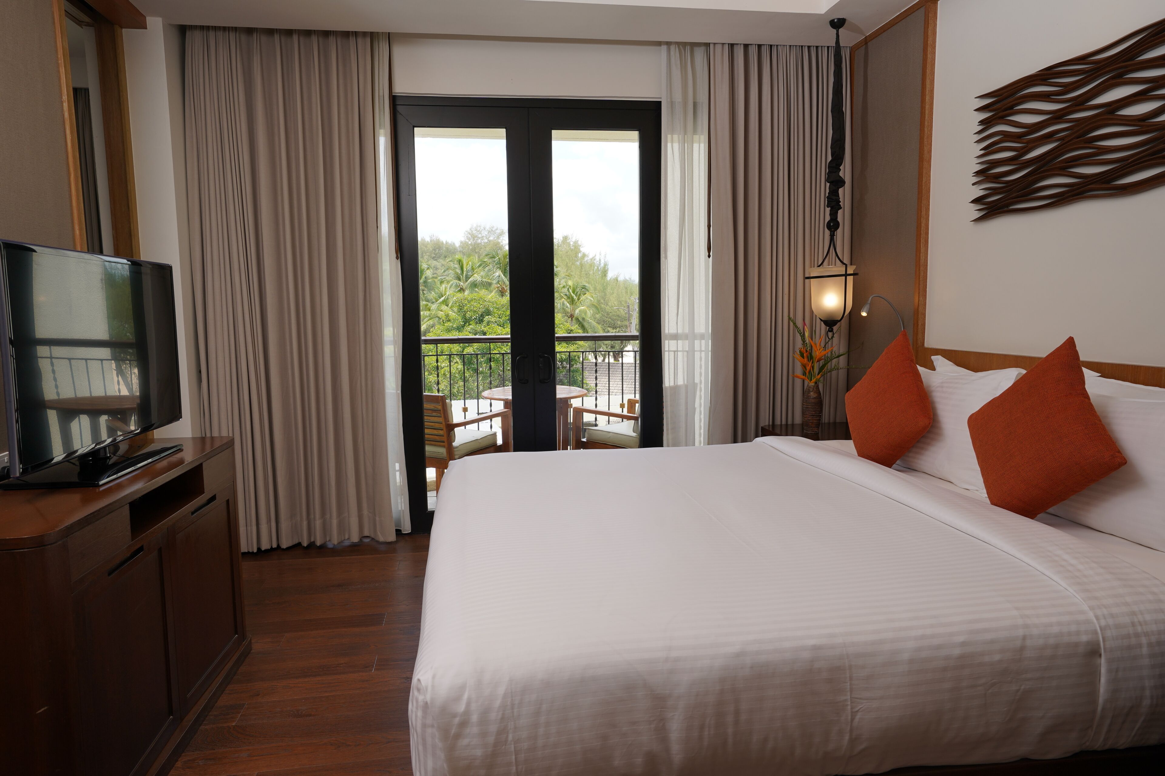 A hotel room with a large bed, a television on a wooden cabinet, a balcony with a view of greenery, and a wall-mounted light fixture.