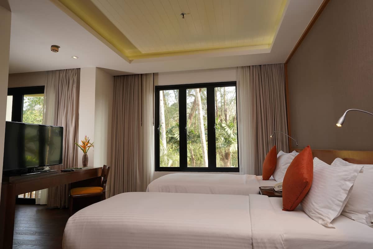 A hotel room with two beds, a desk with a TV, and large windows with curtains.