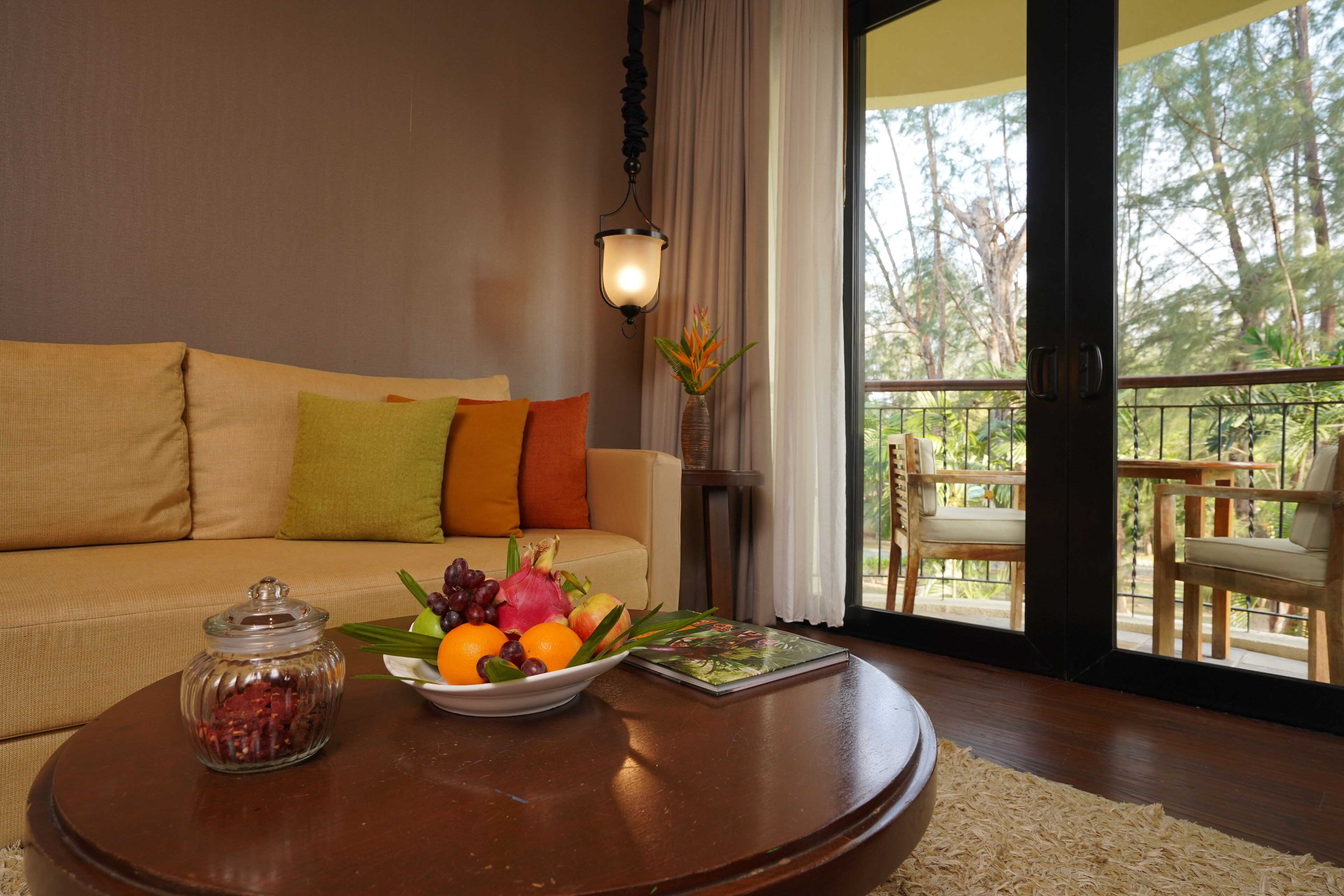 A living room with a sofa, a coffee table with fruit and a book, and a view of a balcony with outdoor furniture.
