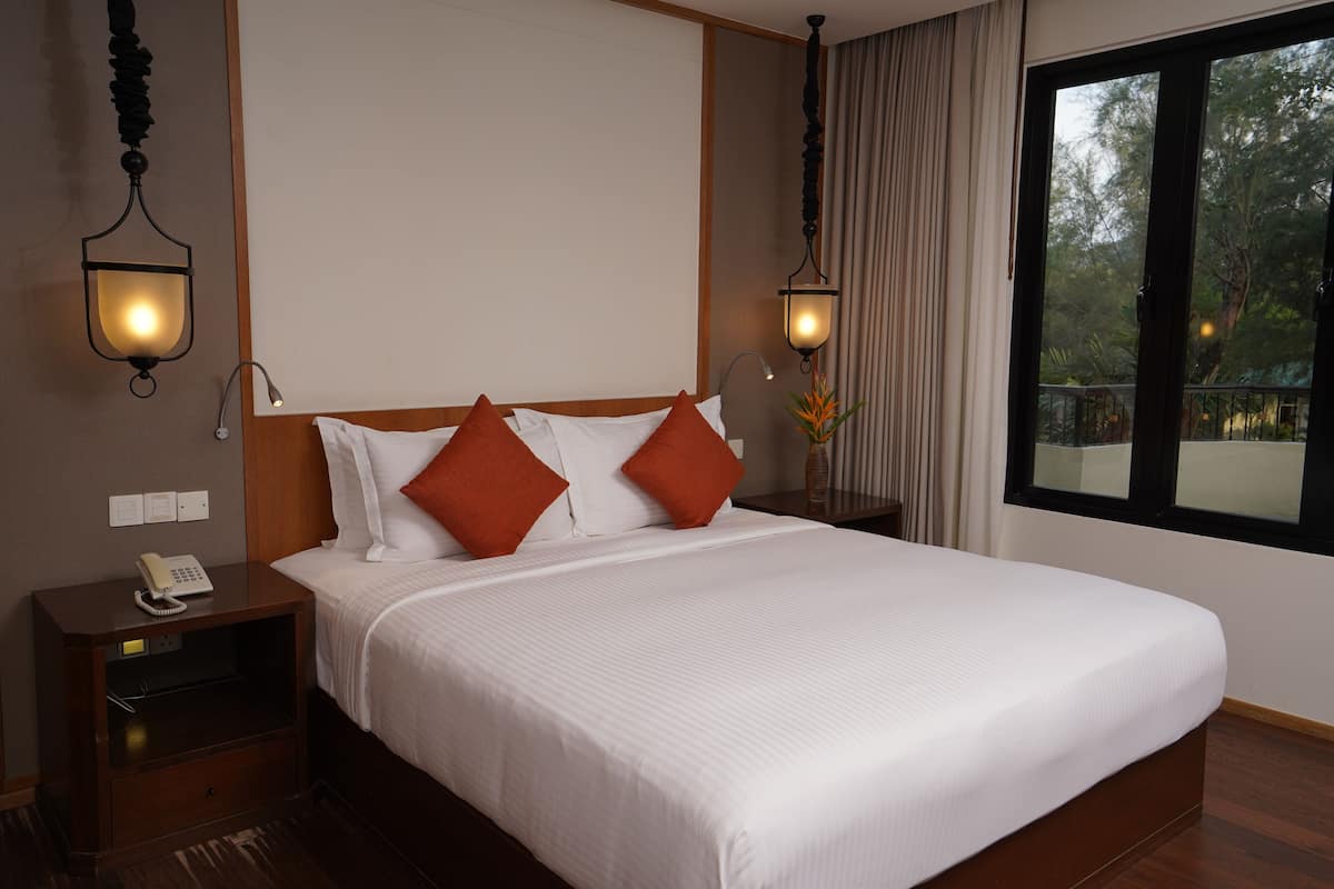 A neatly made bed with white and orange pillows, a bedside table with a lamp, and a view of trees through a window.