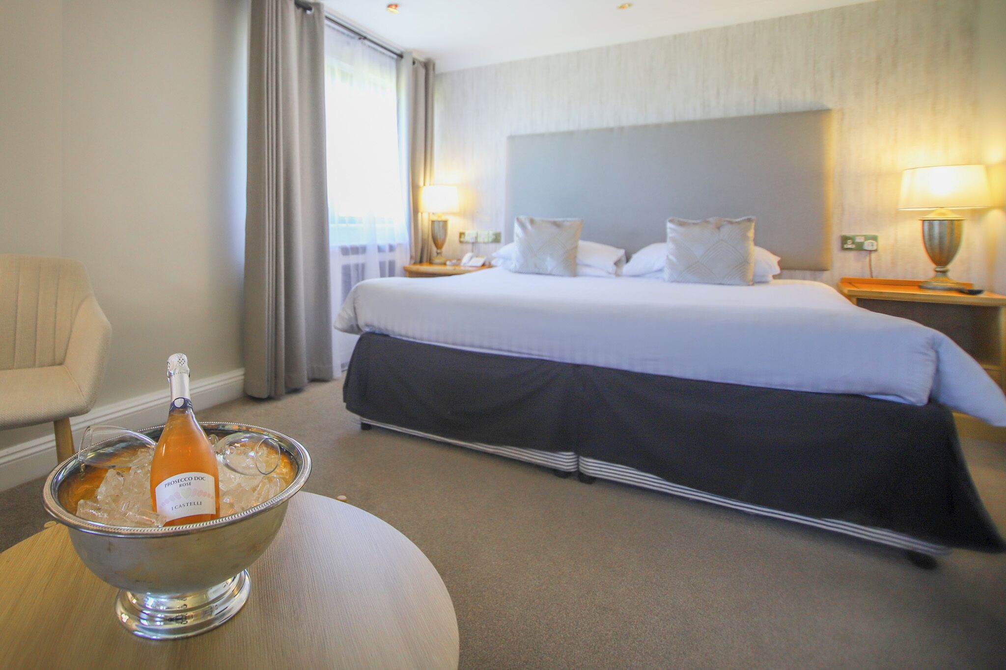 Ein Hotelzimmer mit einem Bett, einem Nachttisch, einer Lampe, einem Sessel und einer Schüssel mit Eis sowie einer Champagnerflasche.