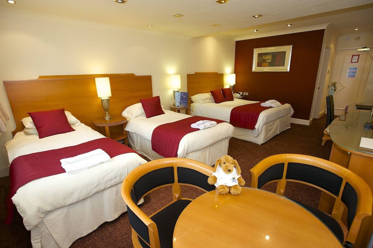 A hotel room with three beds, each with a red blanket and white linens, a wooden headboard, bedside lamps, and a small table with a teddy bear.