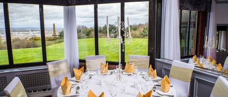 A round dining table set for a formal event with white tablecloths, gold napkins, and silverware. Large windows offer a view of a grassy landscape and a distant monument.