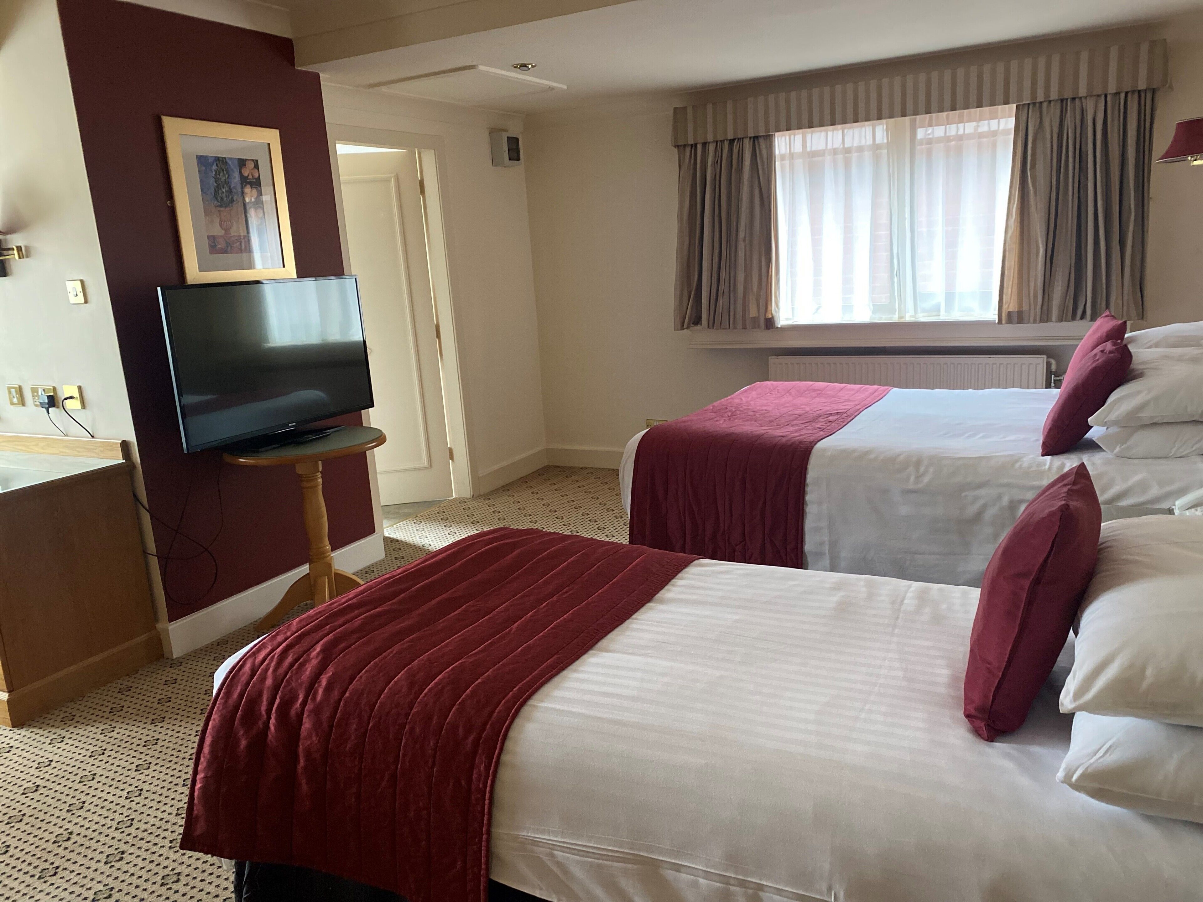 A hotel room with two beds, a television on a stand, a framed picture on the wall, and a window with curtains.