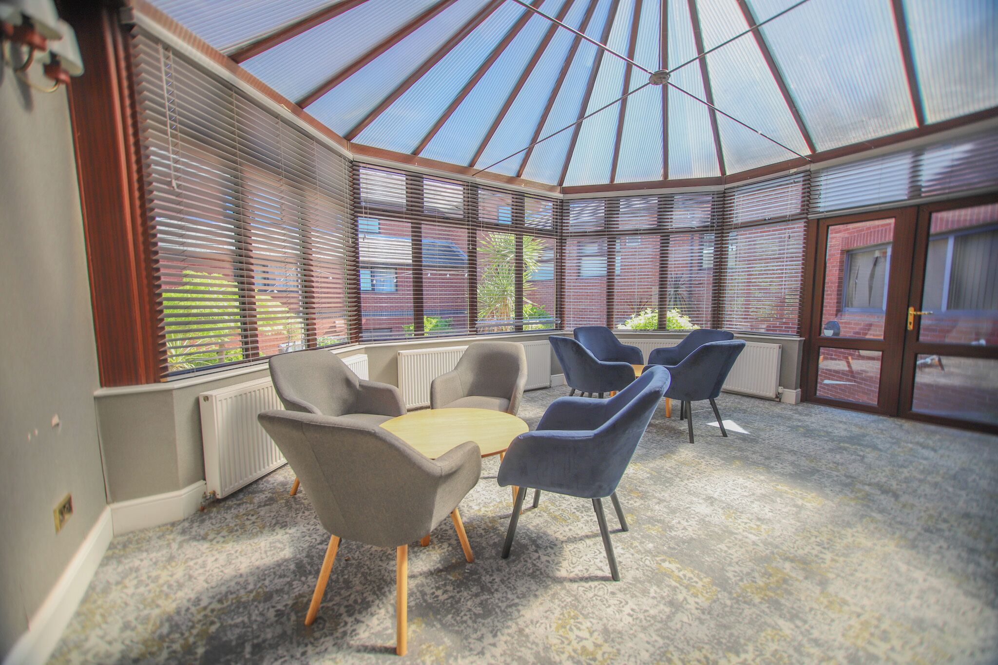 A conservatory with a glass ceiling, seating area with chairs and a table, and a view of the outside.