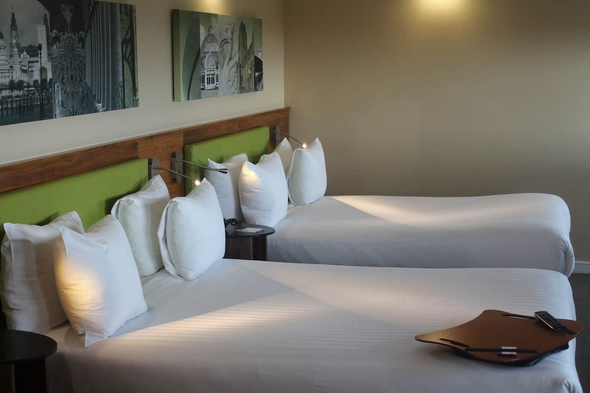 Habitación de hotel con dos camas, cabecera verde, ropa de cama blanca, mesita de noche, control remoto y una obra de arte en la pared.
