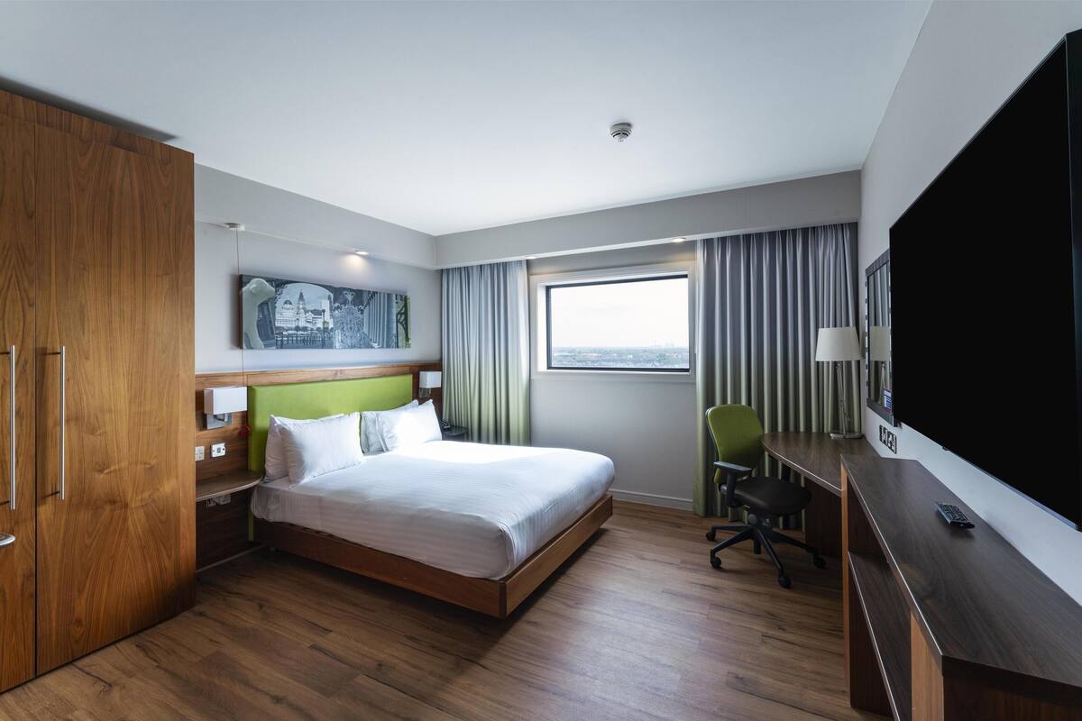 Habitación de hotel con cama, escritorio, televisión, silla y ventana con cortinas.