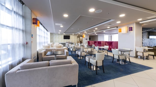 Een moderne hotel lobby met diverse zithoeken, waaronder banken en tafels, en een groot raam met transparante gordijnen.