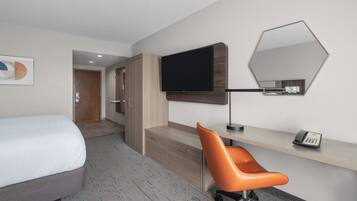 A hotel room with a bed, a desk with a chair, a flat-screen TV, a telephone, and a mirror on the wall.