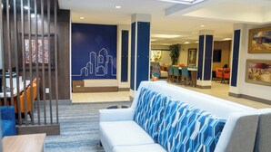 Een moderne hotel lobby met een witte bank, blauw gestreepte kussens, een muurschildering van een stadsgezicht en een eetgedeelte met oranje stoelen.