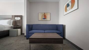 A modern hotel room with a blue sofa, a wooden coffee table, a bed with white bedding, and a framed abstract painting on the wall.
