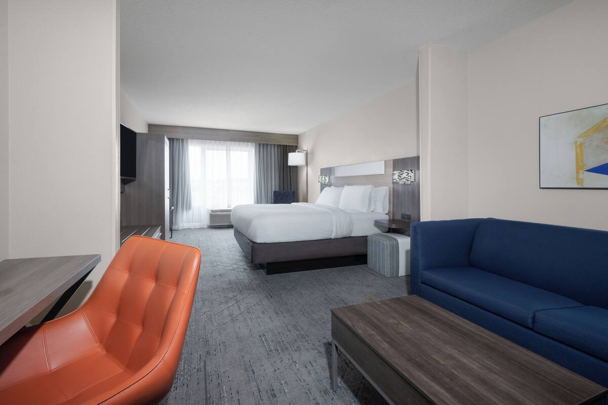 A hotel room with a bed, a blue sofa, an orange chair, a wooden coffee table, and a TV on a stand.