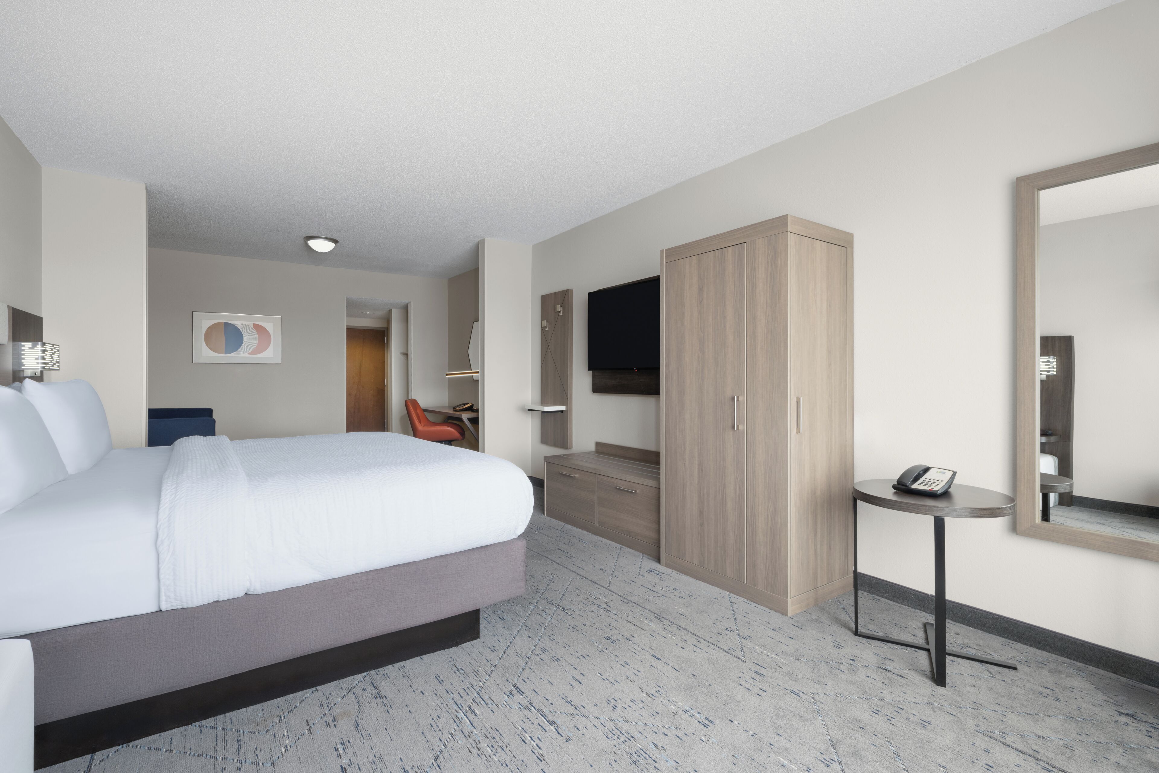 Ein modernes Hotelzimmer mit einem großen Bett, einem Holzschrank, einem kleinen Tisch mit Telefon und einem an der Wand montierten Fernseher.