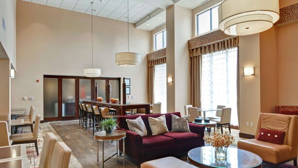 A spacious hotel lobby with a red sofa, leather armchairs, a round glass table, and a dining area with a wooden table and chairs. - Hampton Inn & Suites Peoria at Grand Prairie, IL (Peoria)
