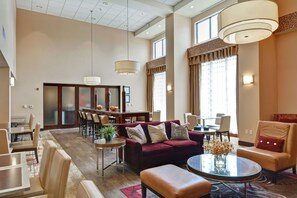 A spacious hotel lobby with a red sofa, leather armchairs, a round glass table, and a dining area with a wooden table and chairs.