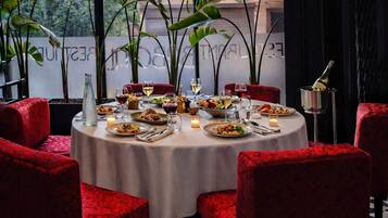 A round dining table with red upholstered chairs, set with plates of food, glasses of wine, and candles.