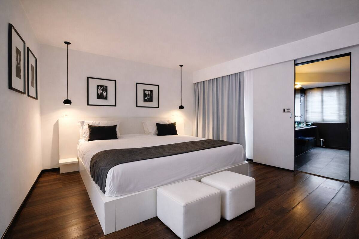 A modern bedroom with a large bed, bedside tables, a bench, framed pictures, and a view into a bathroom.