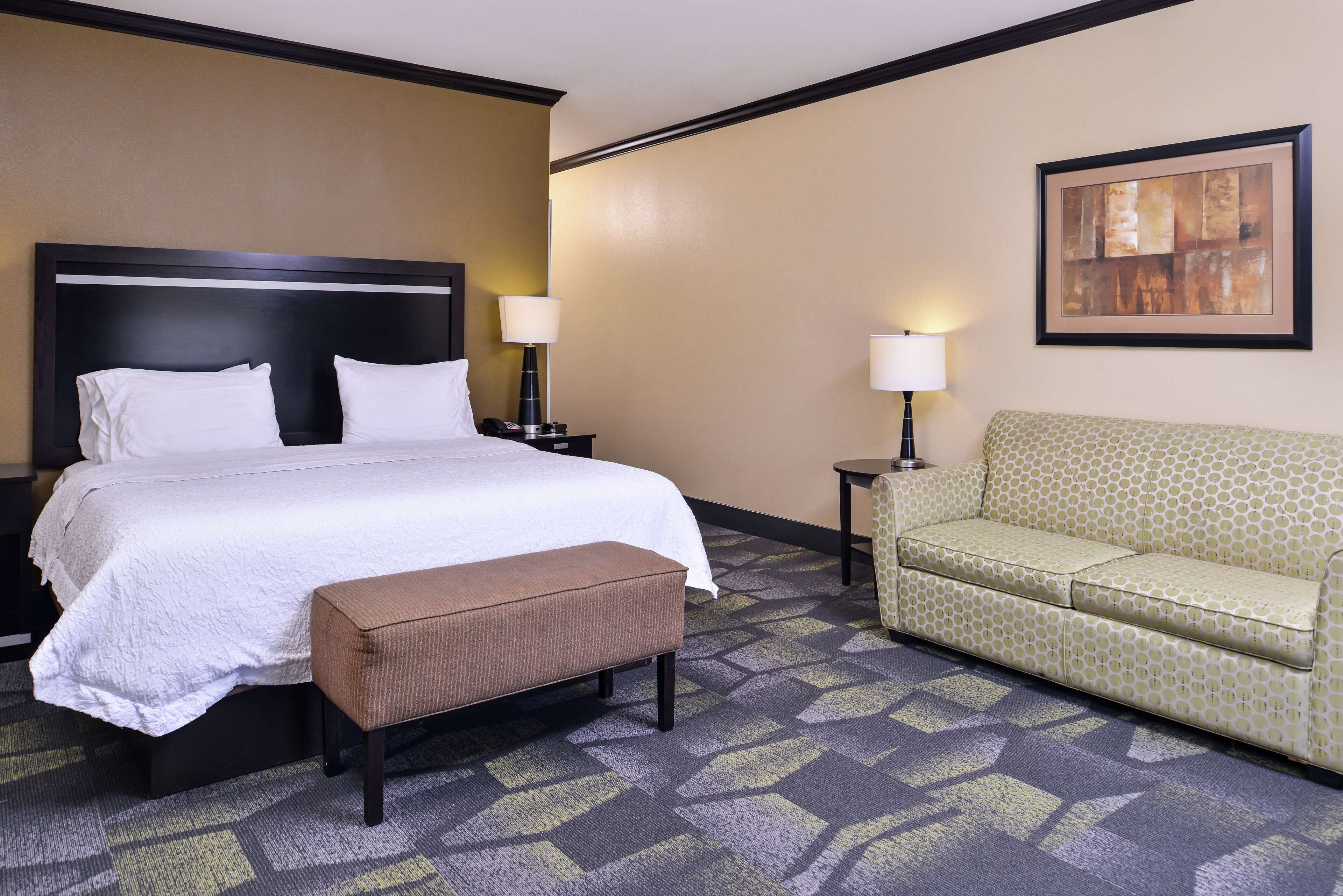 A hotel room with a large bed, a bench, a sofa, two bedside tables, and a wall-mounted lamp.