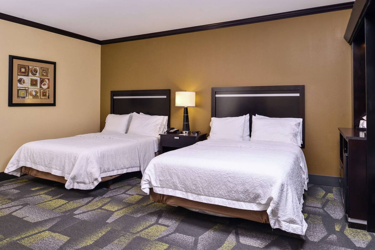 Habitación de hotel con dos camas, una mesita de noche con lámpara y un cuadro en la pared.