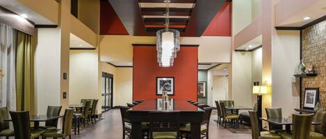 A spacious hotel dining area with a long wooden table surrounded by chairs, a chandelier, and a patterned rug.