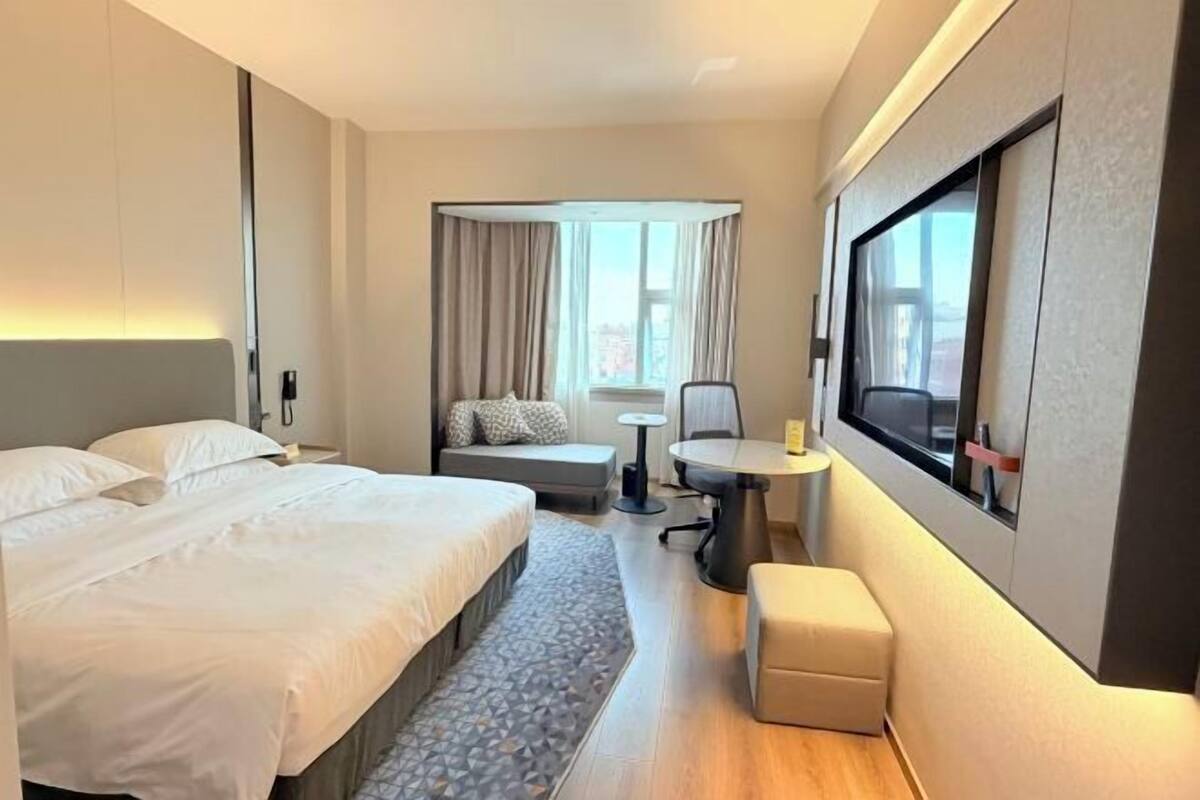 A hotel room with a large bed, a small desk, a chair, a sofa, and a large window with curtains.