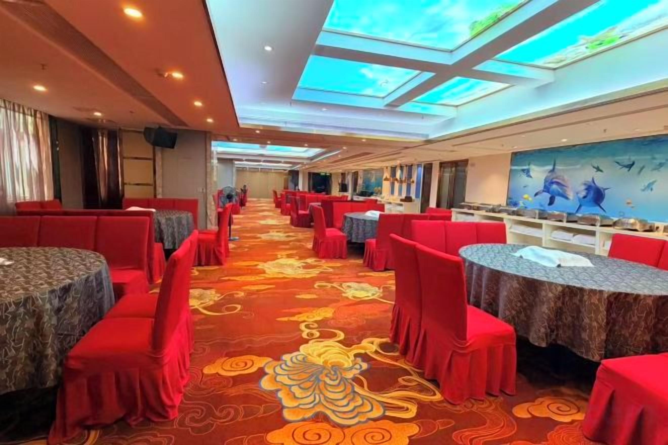 A banquet hall with round tables and red chairs, a patterned carpet, and a ceiling with skylights.