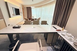 A hotel room with a large bed, a desk with a chair, a dining table with chairs, a TV mounted on the wall, and a view of the city through large windows.