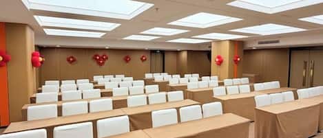 Un salón de conferencias con filas de sillas blancas, mesas cubiertas con manteles marrones y un techo alto con tragaluces.