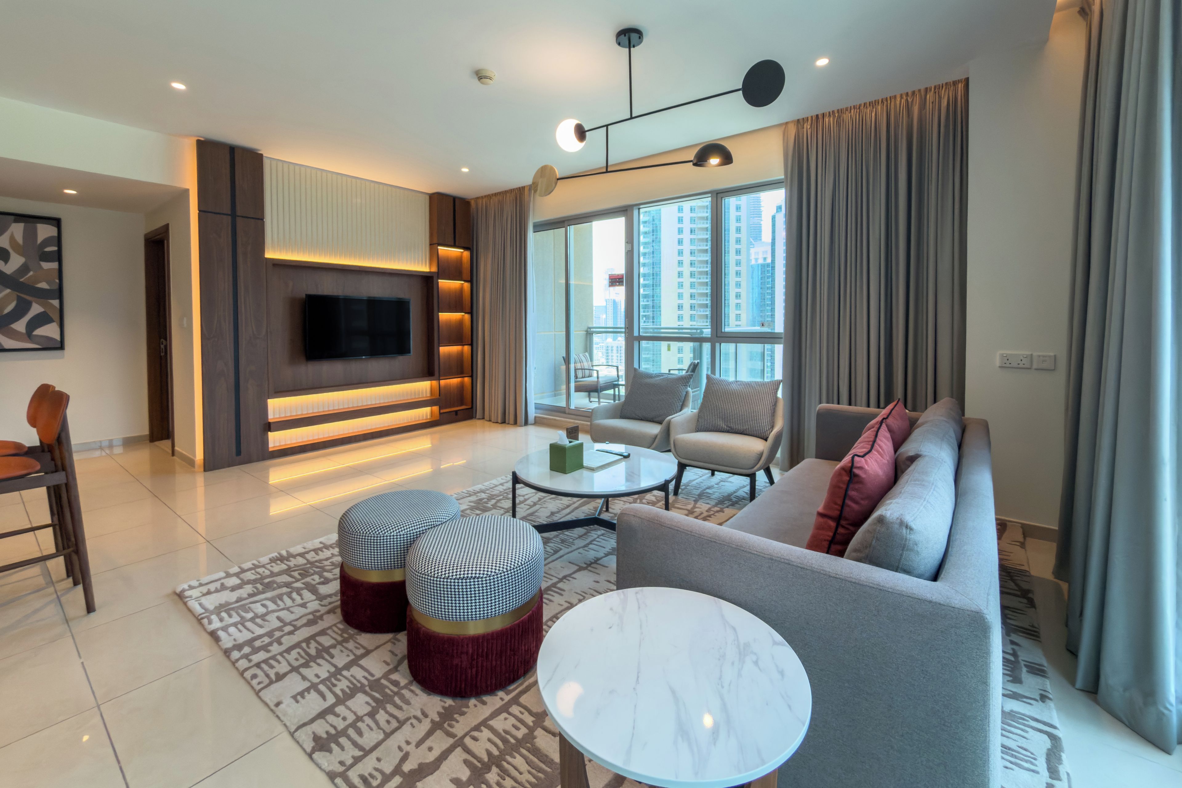 A modern living room with a flat-screen TV, a sofa, two ottomans, a coffee table, and a patterned rug.
