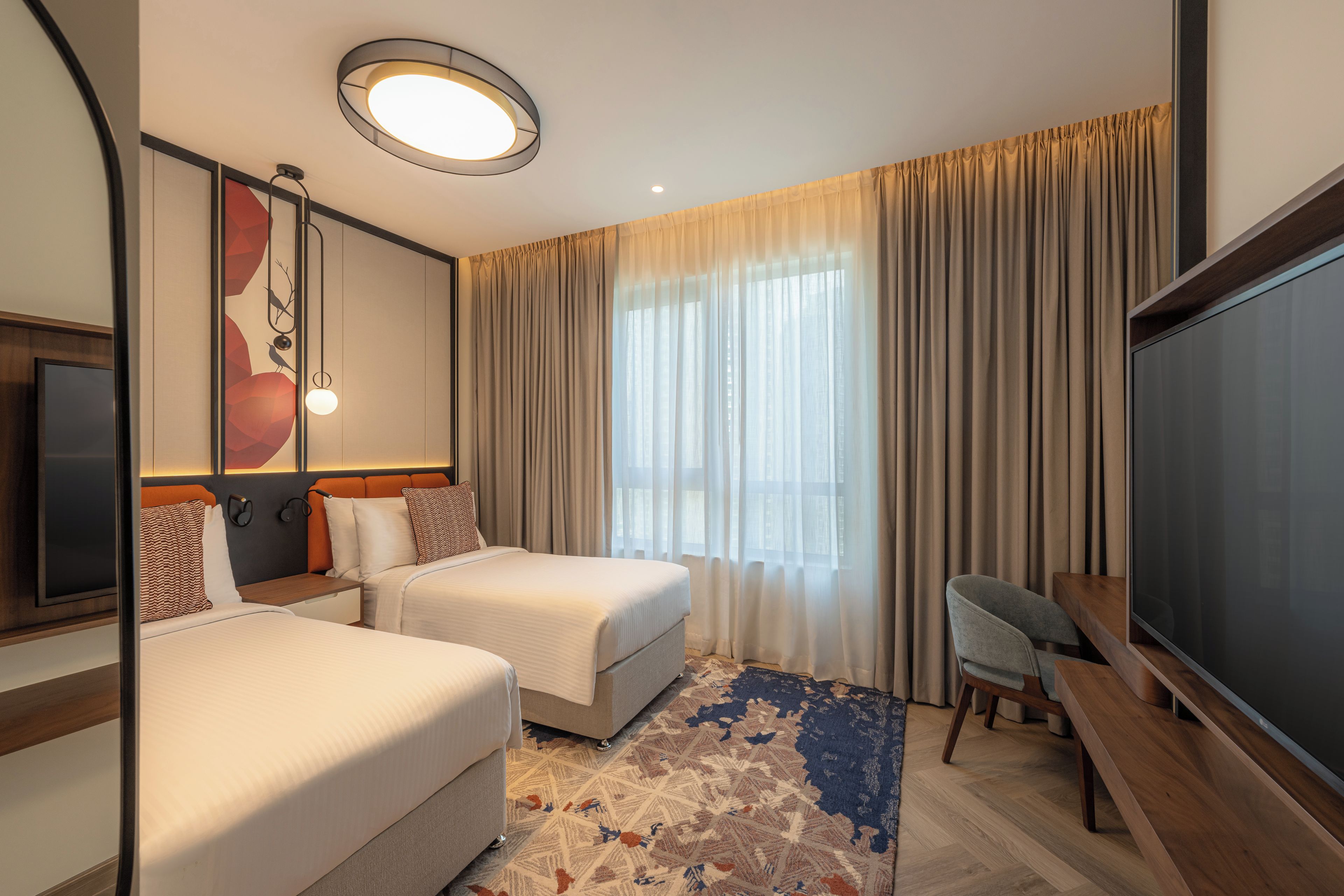 A hotel room with two beds, a desk, a flat-screen TV, a large window with curtains, and a ceiling light.