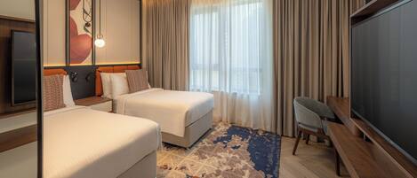 A hotel room with two beds, a desk, a flat-screen TV, a large window with curtains, and a ceiling light.