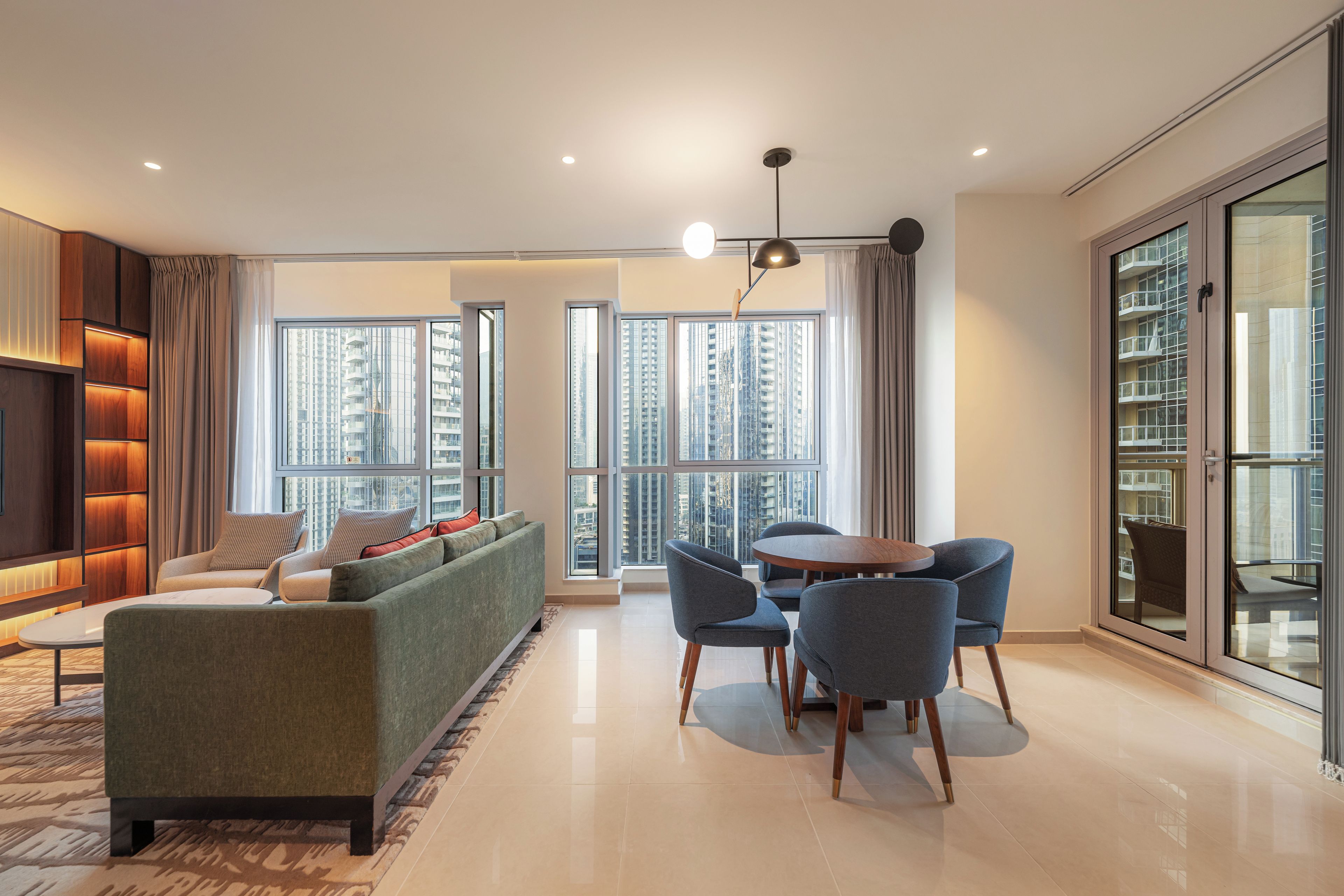 A modern living room with a sofa, chairs, a dining table, and large windows offering a city view.
