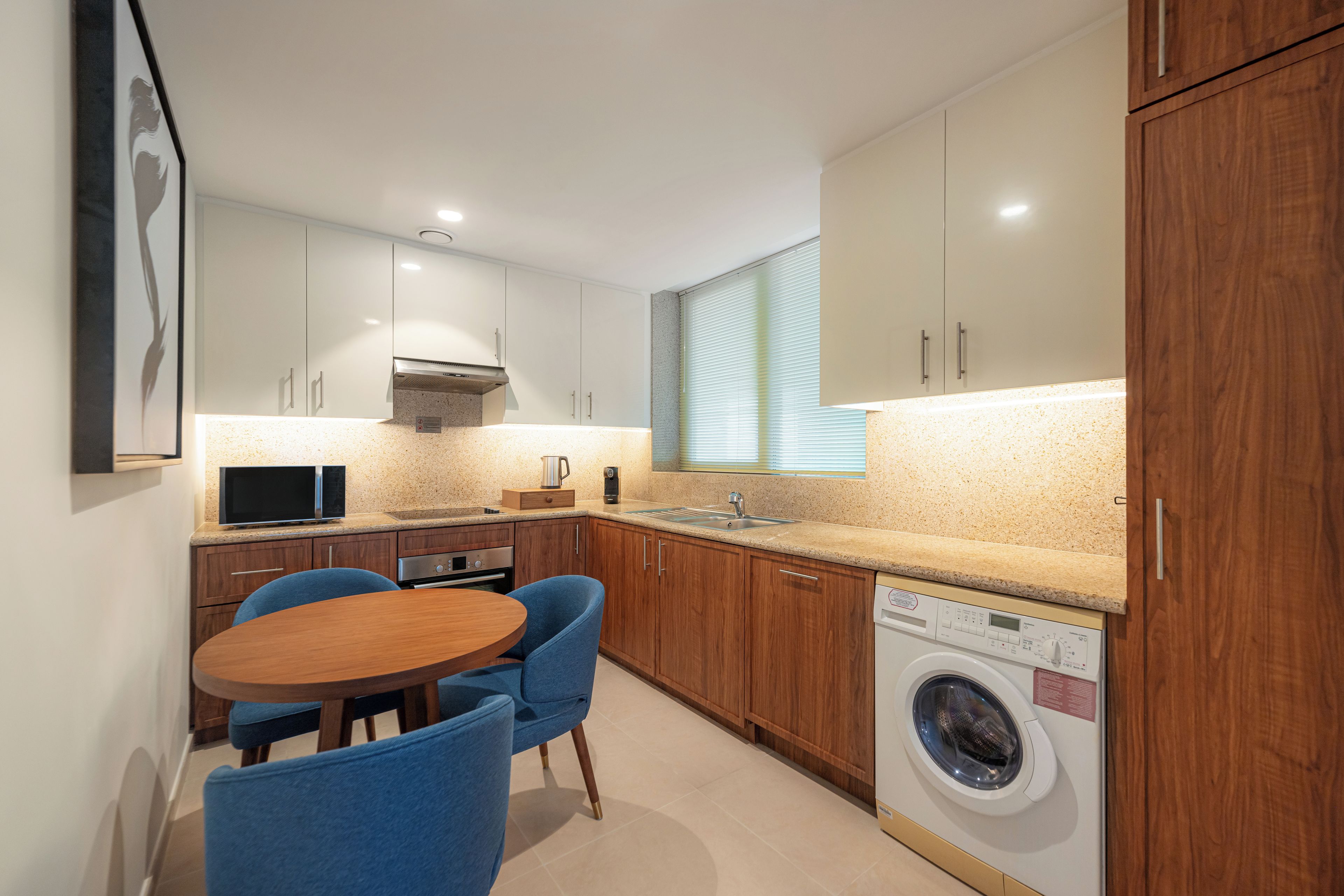 Une cuisine compacte équipée d’un four à micro-ondes, d’un évier et d’un lave-linge, avec des placards en bois et un coin repas comprenant des chaises.