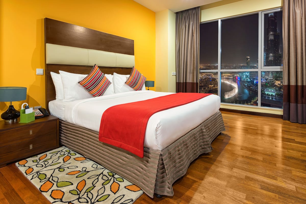 Een hotelkamer met een groot bed, een nachtkastje, een lamp, uitzicht op een stadsbeeld door een raam en een kamerbreed tapijt.