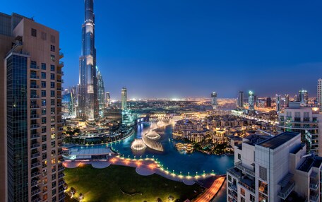 A high-rise building with a distinctive spire, surrounded by a well-lit urban area with a river and a landscaped area with pathways.
